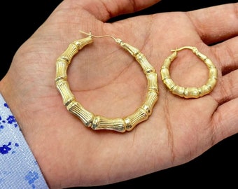 May include: A pair of gold-colored bamboo-style hoop earrings. One large earring and one smaller earring are displayed. The earrings have a textured, ribbed design, and are presented against a neutral background.