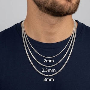 May include: Three silver box chain necklaces of varying thicknesses, 2mm, 2.5mm, and 3mm, are displayed. The chains are worn around a person's neck, showcasing the different sizes. The image is set against a dark blue background.