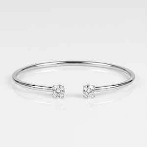 May include: A silver open cuff bracelet featuring two clear, round gemstones at each end. The bracelet is presented on a white surface, reflecting light. A minimalist design.