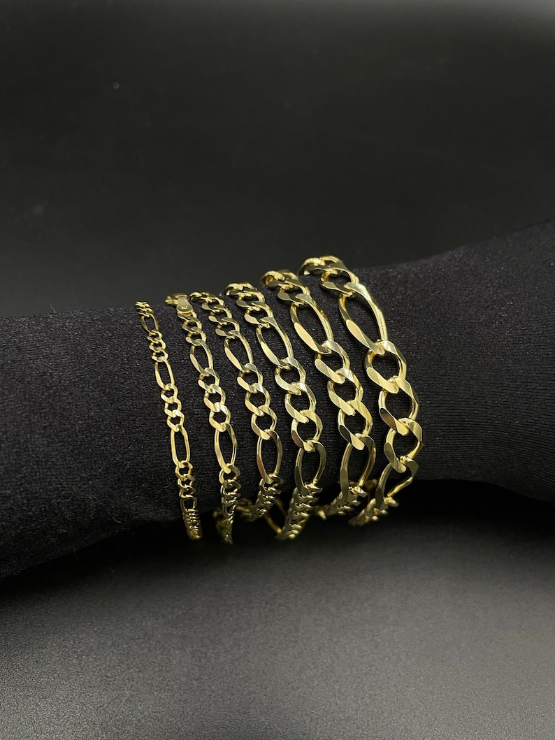May include: A set of six gold chain bracelets of varying widths and styles. The chains are arranged in a stack on a black surface.