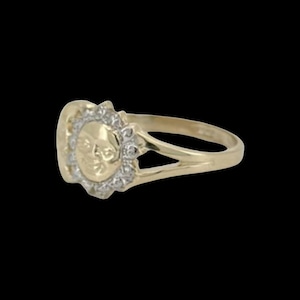 May include: A gold ring with a round face featuring a detailed design of a person's face. The face is surrounded by a circle of small, clear stones.