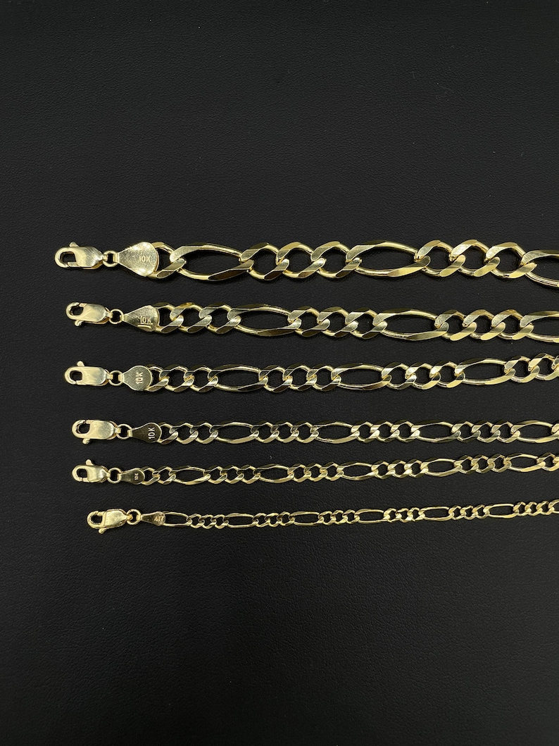 May include: Six gold chains of different lengths and widths, all with a classic figaro design. The chains are laid out in a row on a black background.
