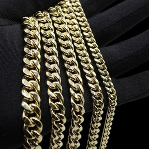 May include: A set of five gold curb chains of varying widths. The chains are laid out in a row on a black surface.