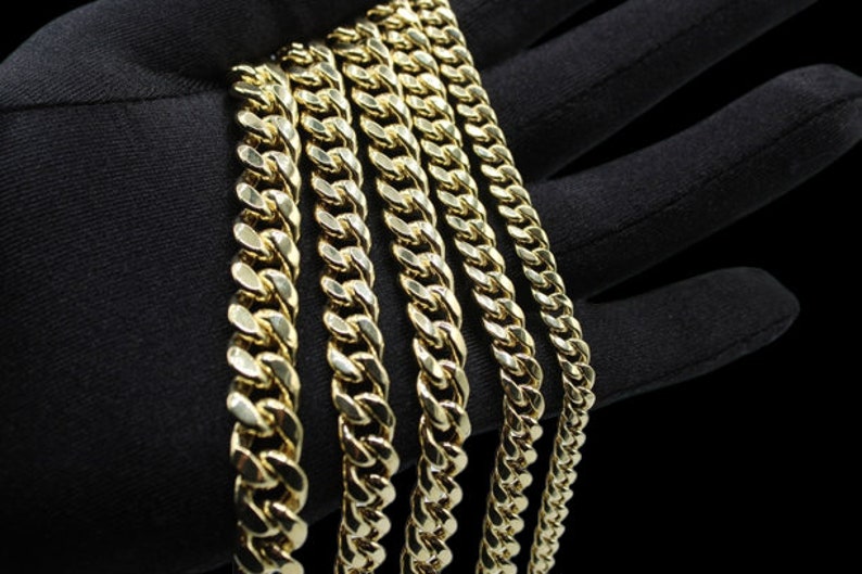 May include: A set of five gold curb chains of varying widths. The chains are laid out in a row on a black surface.