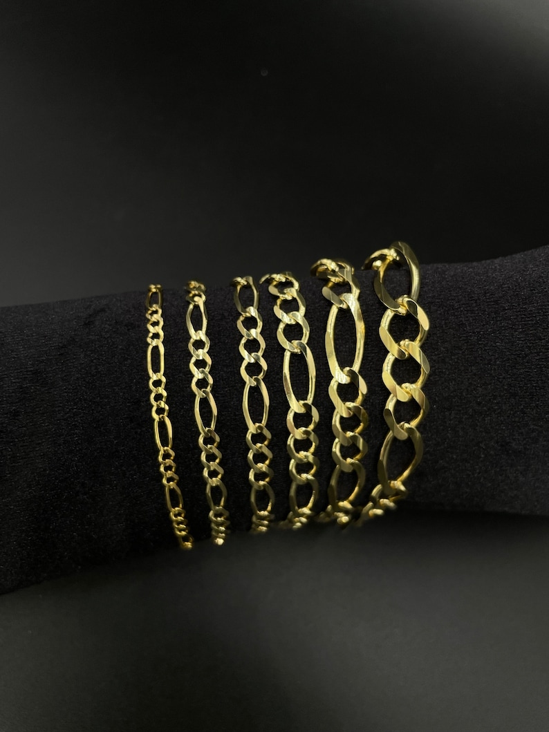 May include: A set of seven gold chain bracelets of varying widths, displayed on a black fabric background.