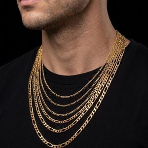 以下が含まれることがあります： さまざまな長さとスタイルのゴールドカラーのチェーンネックレスのコレクション。黒いシャツを着た人物が、黒い背景にネックレスを展示しています。チェーンには、カーブやフィガロスタイルなど、さまざまなリンクデザインがあります。