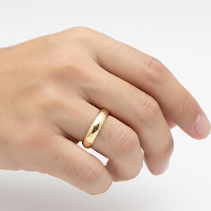 May include: A gold-colored ring with a smooth, rounded design. The ring is shown on a hand, with a bright, reflective surface. The ring appears to be made of a shiny metal, possibly gold or gold-plated.