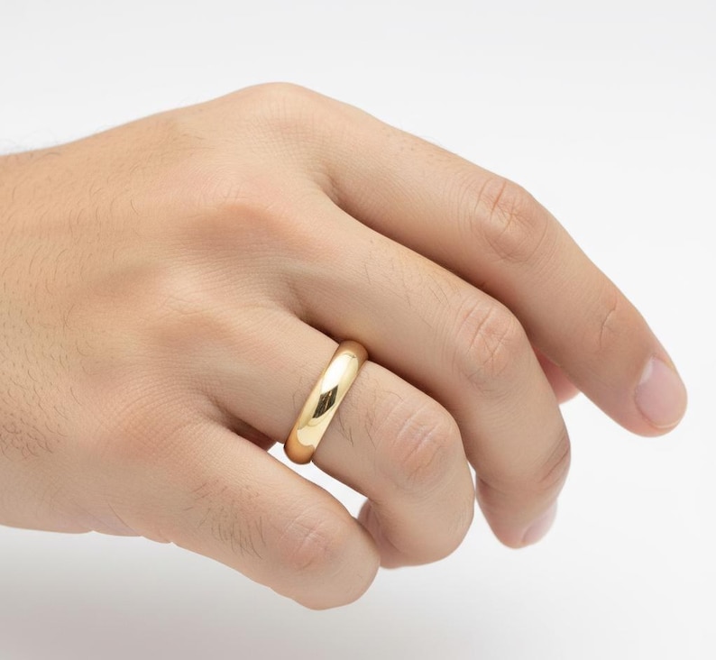 May include: A gold-colored ring with a smooth, rounded design. The ring is shown on a hand, with a bright, reflective surface. The ring appears to be made of a shiny metal, possibly gold or gold-plated.