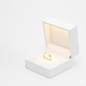 May include: A gold wedding band displayed in an open white ring box with a soft, illuminated interior. The ring is centered on a white cushion, showcasing its simple, elegant design. The box is slightly angled, revealing the ring's polished surface.