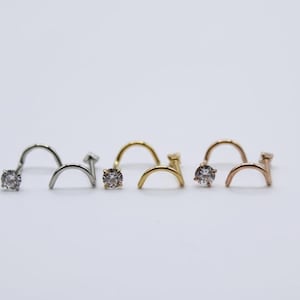 May include: A collection of nose studs in silver, gold, and rose gold tones. Each stud features a small, clear gemstone at the end of a curved post. The jewelry is displayed against a plain white background.