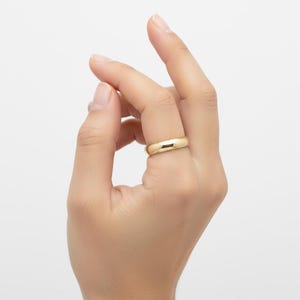 May include: A gold-colored ring with a smooth, rounded band is worn on a finger. The ring has a polished finish and reflects light. The ring is a simple, classic design, suitable for everyday wear.