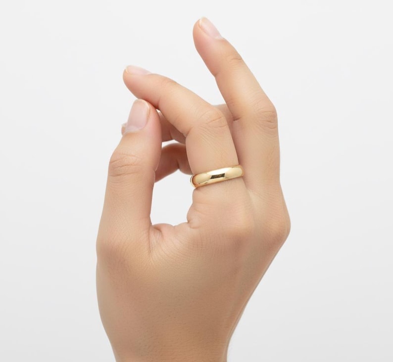 May include: A gold-colored ring with a smooth, rounded band is worn on a finger. The ring has a polished finish and reflects light. The ring is a simple, classic design, suitable for everyday wear.