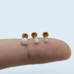 Puede incluir: Se muestran tres pendientes de botón dorados con piedras preciosas de ópalo blanco. Los pendientes son de diferentes tamaños, el más grande a la izquierda y el más pequeño a la derecha. Los pendientes están sobre un fondo blanco.