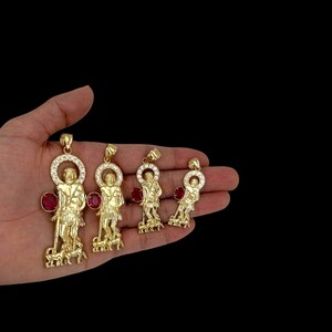 May include: Four gold pendants featuring a religious figure with a red gemstone and a halo of clear stones. The pendants are of different sizes and are displayed on a hand.