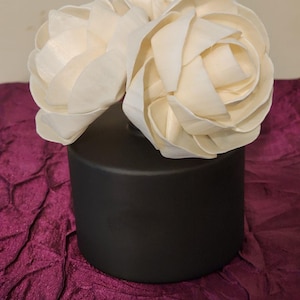 May include: A black diffuser bottle with a white wooden flower diffuser on top. The diffuser is sitting on a purple fabric surface.