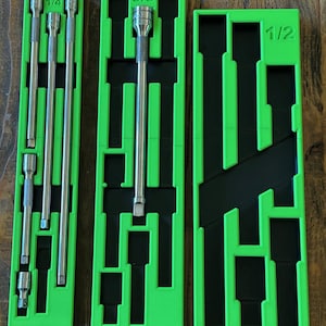 May include: Three green socket organizer trays with black inserts. The trays are labeled 1/4, 3/8, and 1/2, and hold various sizes of silver sockets. The trays are arranged on a wooden surface.