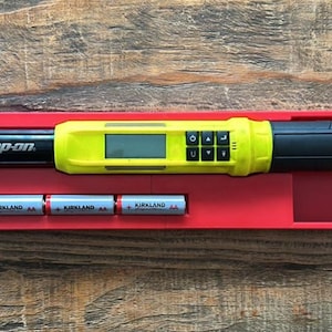 May include: A black and yellow Snap-on digital torque wrench with a digital display and control buttons, resting in a red tray. Three Kirkland batteries are also in the tray. The wrench is on a wooden surface.