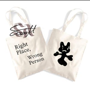 May include: Two white canvas tote bags. One bag has the text "Right Place, Wrong Person" printed on it. The other bag has a black cartoon character with a surprised expression printed on it.
