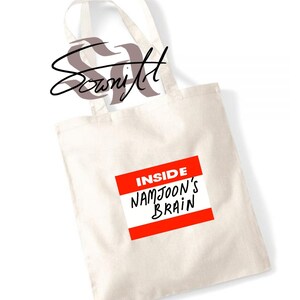 May include: Cream-colored tote bag with black handles. A red and white label on the front reads "INSIDE NAMJOON'S BRAIN" in black lettering. The bag also features a stylized signature in the upper left corner.