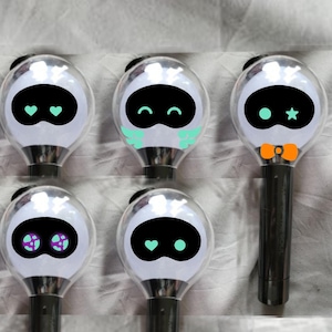May include: A collection of five light sticks with a clear, spherical head and a black handle. Each light stick features a black oval face with different colored accents, including hearts, stars, and a bow tie. The light sticks are designed with a cute, cartoonish aesthetic.