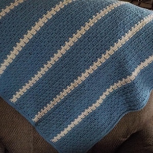 Peut inclure: Une couverture crochetée bleue et blanche avec des rayures diagonales. La couverture est de couleur bleu moyen avec des rayures blanches. La couverture est pliée sur une surface marron. La couverture semble être faite à la main.