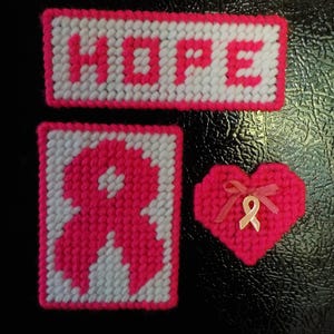 Peut inclure: Trois articles faits à la main : un panneau rectangulaire avec le mot "HOPE", un carré avec un ruban rose et un cœur rose avec un ruban et un petit ruban. Tous les articles sont roses et blancs.