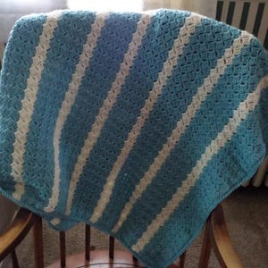 May include: A handmade crochet blanket in shades of blue and cream, draped over a wooden chair. The blanket features a diagonal striped pattern, with alternating bands of light blue and cream-colored yarn. The texture of the crochet stitches is visible.