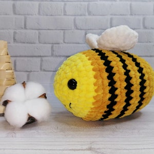 Pode incluir: Uma abelha de pelúcia listrada em amarelo e preto com asas brancas. A abelha tem um rosto sorridente com olhos pretos. Uma flor de algodão e uma cesta de vime estão no fundo.