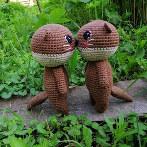 May include: Two handmade crochet otter dolls. The otters have brown bodies, heads, and tails, with cream-colored faces and white whiskers. They have black eyes and pink accents. The dolls are standing on a wooden surface, surrounded by green foliage.