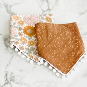 Op de afbeelding: Twee stoffen baby-slabbetjes met witte pomponrand. E&eacute;n slabbetje is bruin met witte stippen en de andere is wit met een bloemenpatroon in roze, gele en groene tinten.