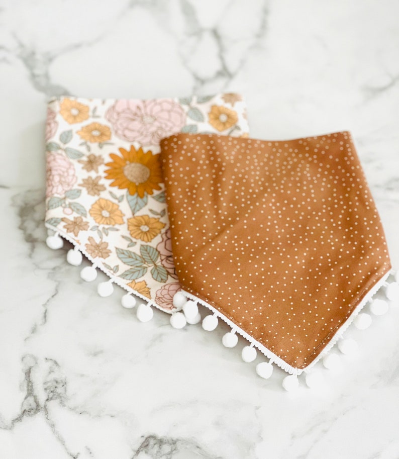 Op de afbeelding: Twee stoffen baby-slabbetjes met witte pomponrand. E&eacute;n slabbetje is bruin met witte stippen en de andere is wit met een bloemenpatroon in roze, gele en groene tinten.