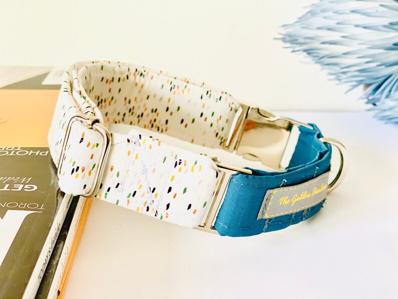 May include: A white dog collar with a colorful confetti pattern and a blue fabric lining. The collar has a silver buckle and a silver D-ring. The collar is labeled "The Golden Buckle".