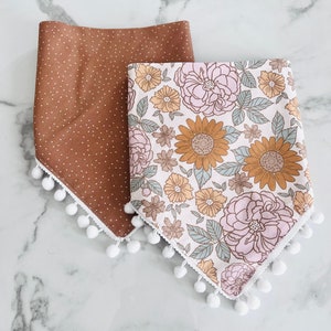 Op de afbeelding: Twee stoffen bandana's in bandana-stijl met witte pomponrand. Een bandana is bruin met witte stippen en de andere is wit met een bloemenpatroon in roze, gele en bruine tinten.