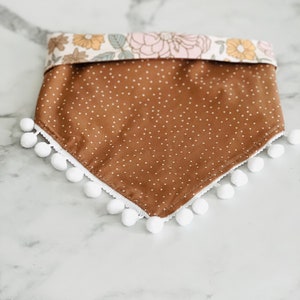 Op de afbeelding: Een bruine bandana met witte stippen en een witte pomponrand. De bandana heeft een bloemenprint aan de bovenrand.