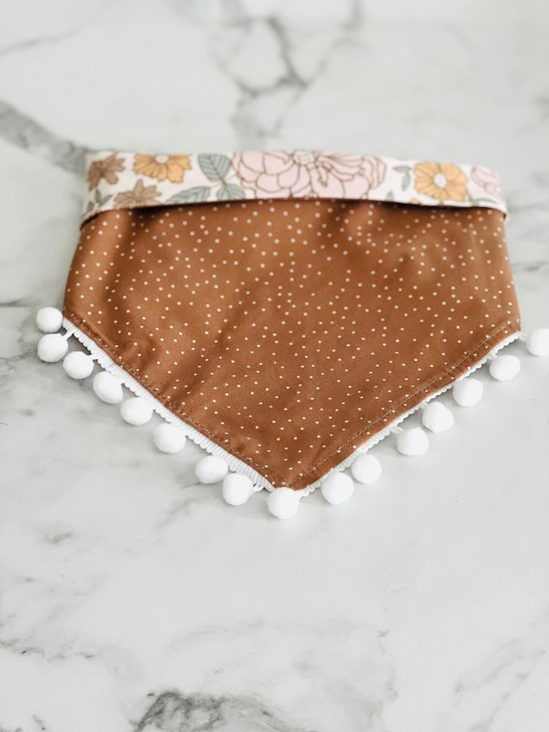 Op de afbeelding: Een bruine bandana met witte stippen en een witte pomponrand. De bandana heeft een bloemenprint aan de bovenrand.