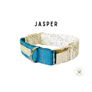 May include: A teal blue dog collar with a white and multi-colored patterned fabric on the inside. The collar has a silver buckle and a yellow label that says "Jasper".