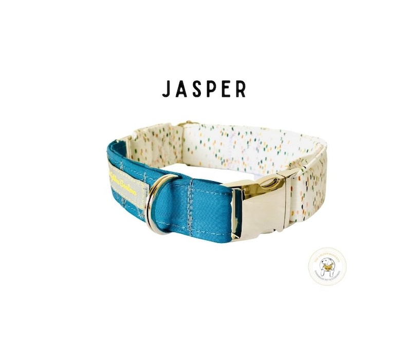 May include: A teal blue dog collar with a white and multi-colored patterned fabric on the inside. The collar has a silver buckle and a yellow label that says "Jasper".