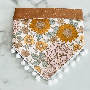 Op de afbeelding: Een witte bandana met een bloemenprint met zonnebloemen, roze bloemen en groene bladeren. De bandana heeft een bruine stippenrand en witte pompoenen rond de randen.