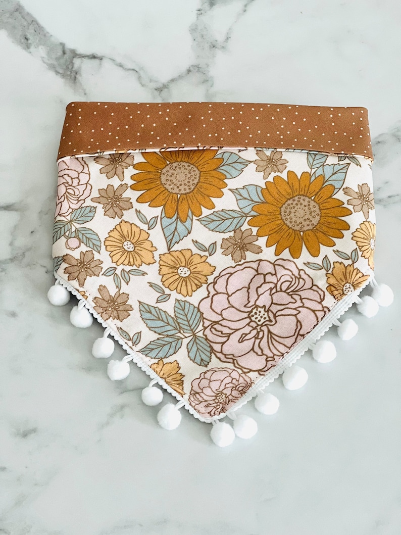 Op de afbeelding: Een witte bandana met een bloemenprint met zonnebloemen, roze bloemen en groene bladeren. De bandana heeft een bruine stippenrand en witte pompoenen rond de randen.