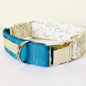 May include: A teal blue dog collar with a white and multi-colored confetti pattern on the other side. The collar has a silver buckle and a silver D-ring.