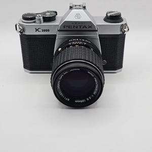May include: A silver and black Asahi Pentax K1000 35mm film camera with a black lens attached. The lens is marked SMC Pentax-M 1:3.5 135mm and Asahi Opt. Co. Japan. The camera body features the K1000 model name and the Asahi Pentax brand logo.
