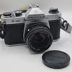 May include: A silver and black Asahi Pentax K1000 35mm film camera with a black SMC Pentax-M 50mm 1:2 lens attached. The camera body features textured black grips, silver metal top and bottom plates, and a separate black circular lens cap with Pentax text.