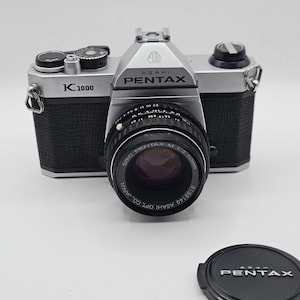 May include: An Asahi Pentax K1000 35mm film camera with a silver and black body, featuring an SMC Pentax-M 1:2 50mm lens. The camera includes a black lens cap with the Pentax logo printed in white, and the camera body displays the K1000 model name.