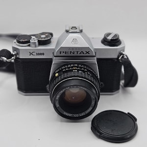 May include: An Asahi Pentax K1000 35mm film camera with a silver and black body. The camera features an SMC Pentax-M 1:2 50mm lens attached to the front, with a black lens cap resting separately on the surface next to the camera body.
