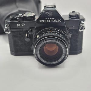 May include: A black Asahi Pentax K2 35mm film camera featuring a textured grip and a mounted SMC Pentax-M 50mm 1:1.7 lens. The camera body displays the K2 model name on the left and the Asahi Pentax brand logo on the top prism housing.