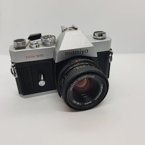 May include: A silver and black Mamiya DSX 1000 35mm film camera featuring a Pentacon auto 1.8/50 multi-coating lens. The camera body includes a top-mounted shutter speed dial, a film advance lever, and a hot shoe for external flash attachments.