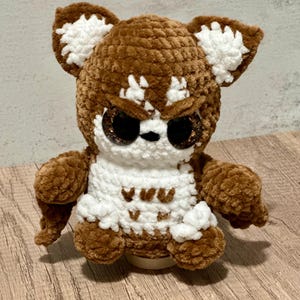 Può includere: Un peluche a forma di gufo fatto a mano, marrone e bianco, con grandi occhi glitterati. Il gufo ha un'espressione severa ed è seduto su una superficie di legno. Il peluche ha accenti bianchi sulle orecchie, sul petto e sui piedi.