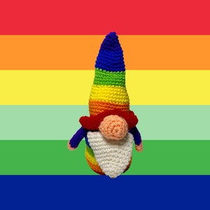 Puede incluir: Un gnomo de crochet con un sombrero de colores arcoíris y barba blanca. El gnomo está de pie frente a un fondo de arcoíris.