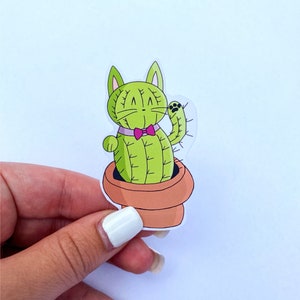 May include: A green cactus shaped like a cat wearing a pink bow tie in a terracotta pot. The cactus is waving with its arm.