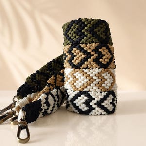 May include: A stack of four macrame straps featuring geometric heart patterns in olive green, tan, cream, and black. Each strap is finished with a bronze-toned metal swivel lobster clasp at the end, designed for attachment to bags or other accessories.